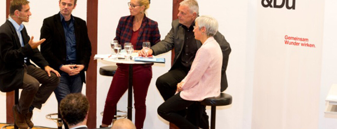 Fünf Menschen bei einem Stehtisch diskutieren miteinander. Im Hintergrund ist ein Plakat der Caritas zu sehen. 