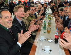 Ca. 10 Menschen sitzen an einem langen Biertisch. Sowohl die junge als auch die alte Generation ist vertreten.  Sie alle klatschen enthisiastisch. 