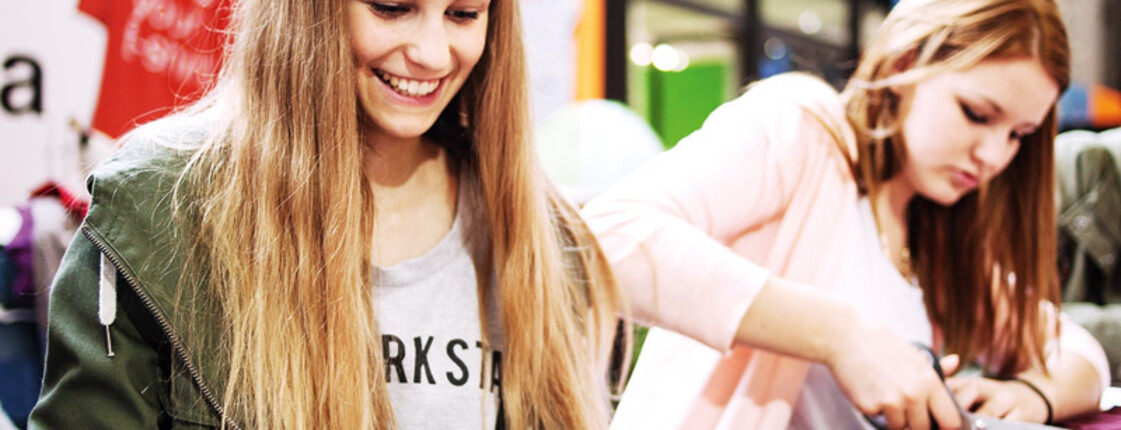 Zwei weibliche Jugendliche mit langen Haaren reparieren Kleidung und gestalten diese mit Scheeren Sie wirken konzentirert und haben Freude an ihrer Tätigkeit. Selber tragen sie ein rosa Oberteil bzw. eine dunkelgrüne Jacke. 