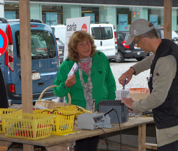 Die carla Reparaturcafés haben in der felkircher Innenstadt einen Stand, bei dem sie Passanten erklärt, wie Geräte repariert werden. Eine Passantin höheren Alters mit grüner Jacke hört dem carla Mitarbeiter aufmerksam zu. 