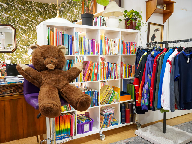 Ein Bereich des carla Shops im Burgenland mit einem großen braunen Plüschbären, einem Regal voller bunter Bücher und einem Kleiderständer mit Secondhand-Kleidung.