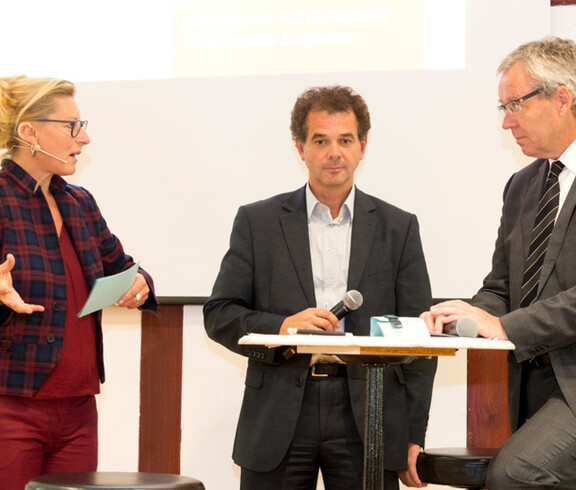 Drei Menschen bei einer Diskussionsrunde. Ein Stehtisch in der Mitte, um den sich zwei Männer befinden. Sie halten jeweils ein Mikrofon in der Hand.