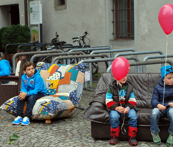 Vier Buben sitzen auf carla Möbeln in der feldkircher Innenstadt am Reuse Day. Es sind Sofas und Sessel. Zwei der Jungen haben jeweils einen roten Luftballon an der Schnur. 