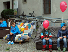Vier Buben sitzen auf carla Möbeln in der feldkircher Innenstadt am Reuse Day. Es sind Sofas und Sessel. Zwei der Jungen haben jeweils einen roten Luftballon an der Schnur. 