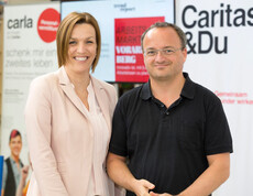 Auf dem Bild ist eine Frau mittleren Alters und ein Herr zu sehen. Beide grinsen. Im Hintergrund sind Plakate der Carla und Caritas.
