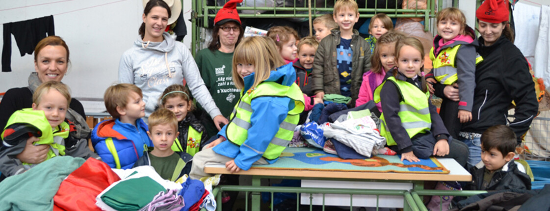 Kindergartenkinder sind zu Besuch beim carla Sortierwerk. Sie stehen neben bzw. sitzen auf den Wägen, in dem sich das sortierte befindet. Manche Kinder tragen eine gelbe Warnweste. 