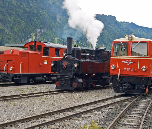 Ein gewöhnlicher Bahnhof mit einer alten, schwarzen Dampflockomotive. Auf den Gleisen daneben stehen zwei rote, etwas modernere Loks. 