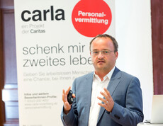 Ein Herr mittleren Alters gestikuliert mit seinen Händen. Er trägt ein Headset sowie eine Brille. Im Hintergrund ist ein Carla Plakat zu sehen.