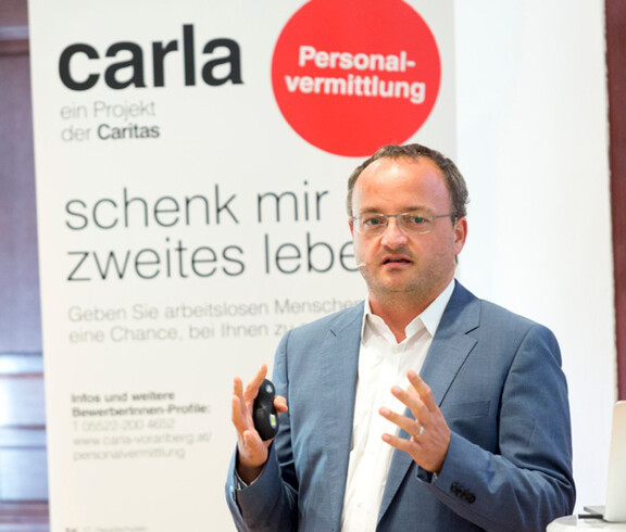 Ein Herr mittleren Alters gestikuliert mit seinen Händen. Er trägt ein Headset sowie eine Brille. Im Hintergrund ist ein Carla Plakat zu sehen.