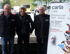 Drei carla Mitarbeiter vom Reparaturcafe posieren zu einem gemeinsamen Gruppenfoto in der feldkicher Innenstadt. Neben ihnen steht ein weißes carla Roll-Up. Die Herren mittleren Alters sind größtenteils schwarz gekleidet. Auf ihren schwarzen Weste steht in Weiß carla. 