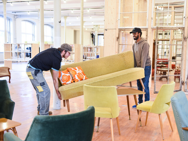 Zwei Mitarbeiter des carla Stores Klarenbrunnen in Bludenz transportieren ein gelbes Sofa in einem großen Raum, der mit weiteren Möbeln ausgestattet ist.