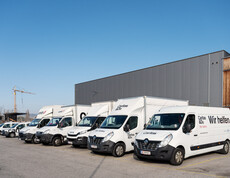 Eine Reihe weißer carla-Transporter steht vor einem Gebäude bereit für den Einsatz. Die Fahrzeuge sind Teil des Logistik- und Serviceangebots von carla.