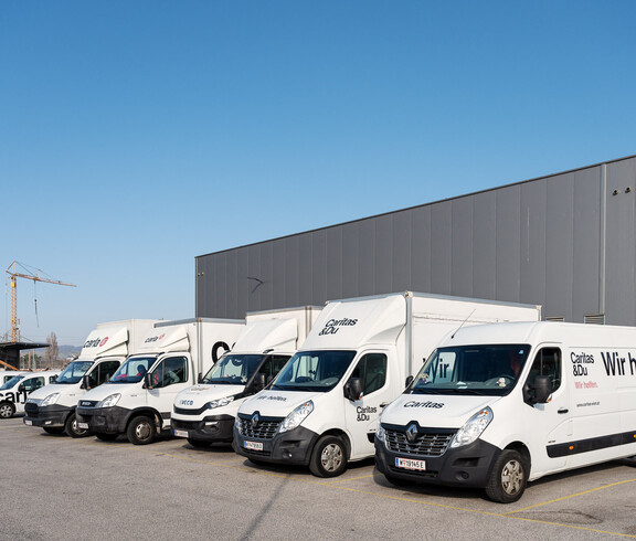 Eine Reihe weißer carla-Transporter steht vor einem Gebäude bereit für den Einsatz. Die Fahrzeuge sind Teil des Logistik- und Serviceangebots von carla.