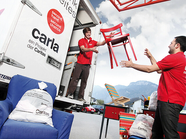 zwei männliche Personen mit roten Tshirts verladen einen roten Stuhl. Der Lastwagen hat die Aufschrift carla.