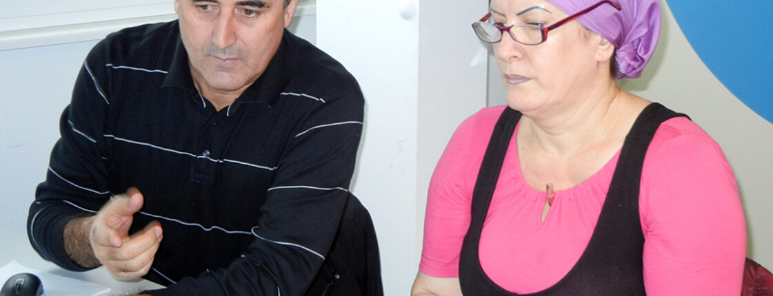 Zwei Flüchtlinge, ein Mann und eine Frau, sitzen an einem Tisch und sind sich am Unterhalten. Der Mann trägt einen schwarzen Pullover, die Frau ein schwarz-rosa Oberteil mit einem violetten Kopftuch. 