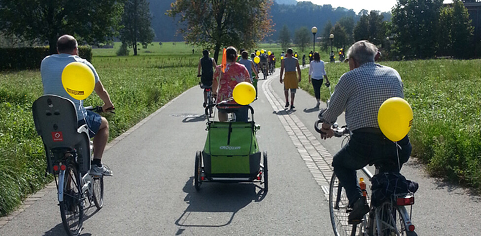 Ca. 10 sich im Bild befindende Personen fahren auf einer flachen Feldstraße Fahrrad. Dabei hat jeder einen gelben Luftballon am Fahrrad angebracht. 