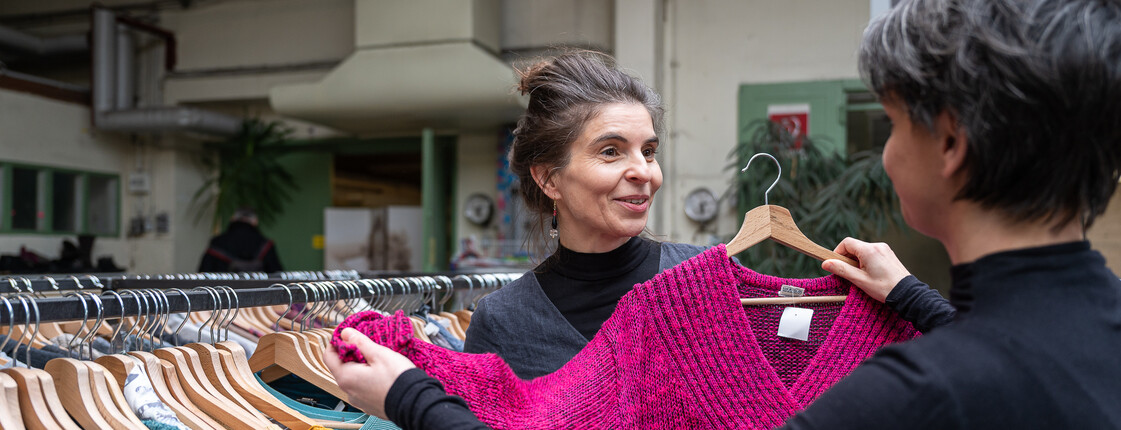 Zwei Frauen in einem Second-Hand-Shop von carla begutachten ein pinkfarbenes Kleidungsstück und lächeln einander zu.