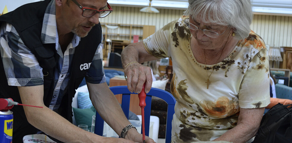 Ein Mann mittleren Alters, der wenige Haare auf dem Kopf hat, und eine ältere Frau mit grauen Haaren reparieren ein kleines, rundes, beiges Elektrogerät auf einem kleinen Tisch. 
