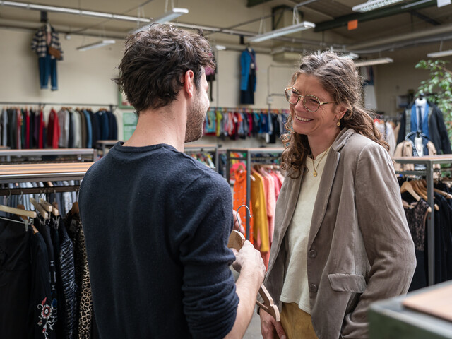 Zwei Menschen im Gespräch und lächelnd in einem Secondhand-Laden, umgeben von bunten Kleiderständern, die eine einladende Einkaufsatmosphäre vermitteln.