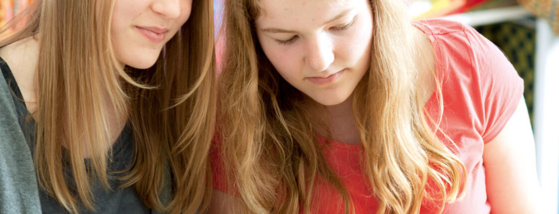 Zwei Mädchen mit langen blonden Haaren sind gerade am Nähen. Das eine trägt ein grünes Obertreil, das andere ein rotes. 