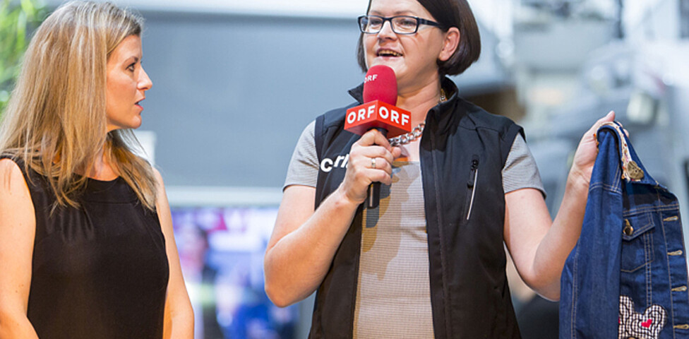 Zwei Frauen mittleren Alters bei einem Interview. Eine Frau hält ein ORF Mikrofon in der Hand und hält eine Jeansjacke in die Höhe. 