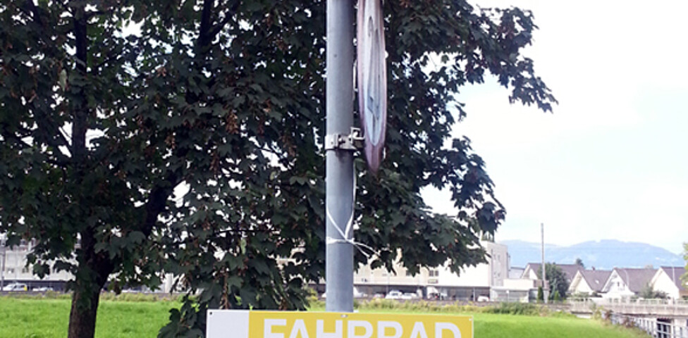 Ein gelb-weißes Schild auf einem metallernem Pfosten wurde angebracht. Darauf zu lesen: Fahrradparade. Auch hängen am Pfosten gelbe Luftballons. 
