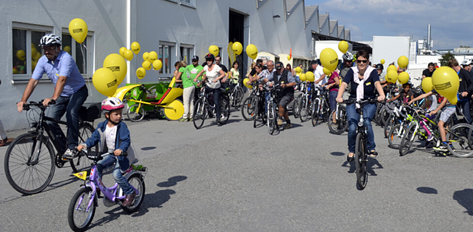 Ca. 20 Menschen mit an ihren Rädern gelb befestigten Luftballonen befinden sich auf einem großen asphaltierten Platz. 