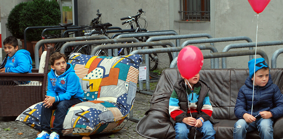 Vier Buben sitzen auf carla Möbeln in der feldkircher Innenstadt am Reuse Day. Es sind Sofas und Sessel. Zwei der Jungen haben jeweils einen roten Luftballon an der Schnur. 