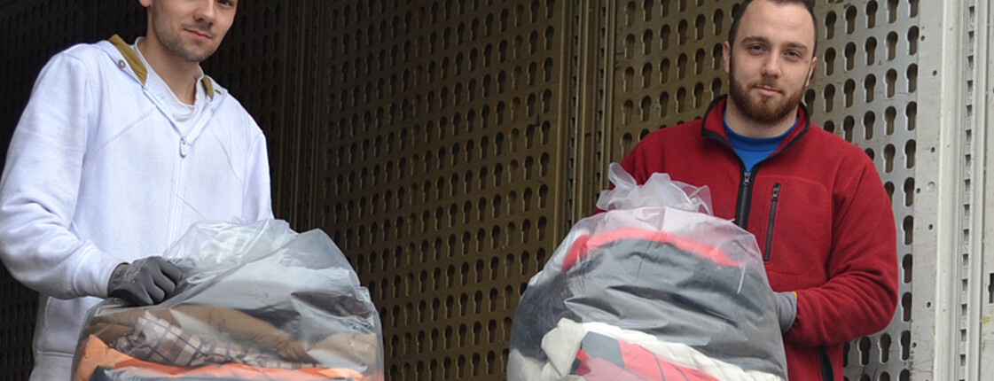 Zwei junge Männer stehen in einem Lastwagen und halten durchsichtige Kleidersäcke in der Hand.