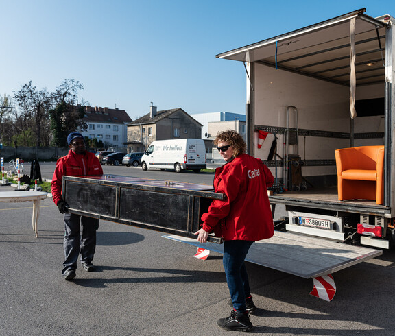 Zwei carla-Mitarbeiter laden gemeinsam ein großes Möbelstück in einen offenen Transporter. Ein orangefarbener Sessel steht bereits im Fahrzeug.