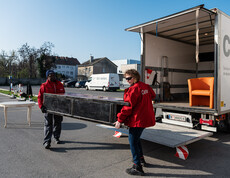 Zwei carla-Mitarbeiter laden gemeinsam ein großes Möbelstück in einen offenen Transporter. Ein orangefarbener Sessel steht bereits im Fahrzeug.