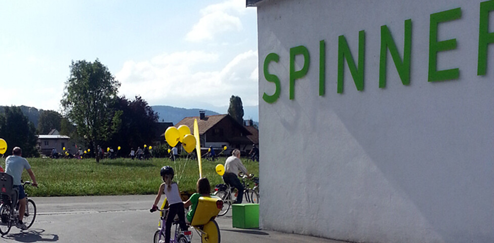Ein Kind fährt mit einem Fahrrad auf Asphalt. Ans Rad wurde ein gelber Luftballon gebunden. Rechts zwei Meter daneben steht eine weiße Wand, auf der "Die Spinnerei" mit grünen Buchstaben steht. 