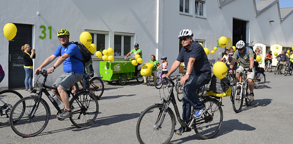 Ca. 10 Menschen mit an ihren Rädern gelb befestigten Luftballonen befinden sich auf einem asphaltierten Platz. 